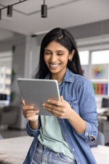 Cheerful young Indian company worker woman in casual using digital tablet for online communication,...