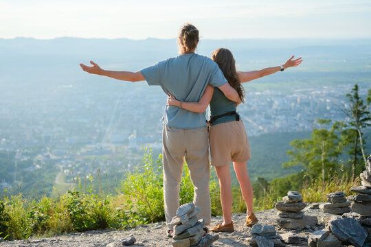 Happy young travelers man and woman standing back view hugging with arms outstretched, admiring beautiful cityscape from mountain viewpoint during their journey. - Powered by Adobe