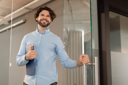 Portrait of confident smiling middle aged business man in shirt opening glass door walking in modern office and smiling, holding clipboard and handle, free copy space, low angle view - Powered by Adobe
