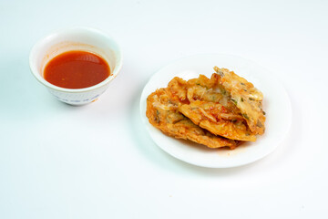 
Description:
A stack of golden-brown bakwan udang, an Indonesian shrimp fritter, sits on a white plate next to a small bowl of sweet chili dipping sauce, a popular street food snack.