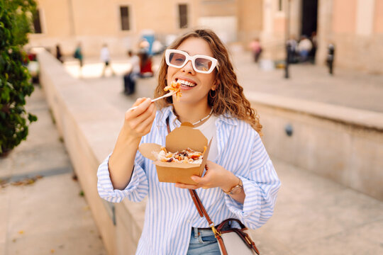 Fototapeta Smiling tourist eats street food from a takeaway box on sunny city streets. Young woman tries Italian pasta in the open air. Food concept, urban, tourism.