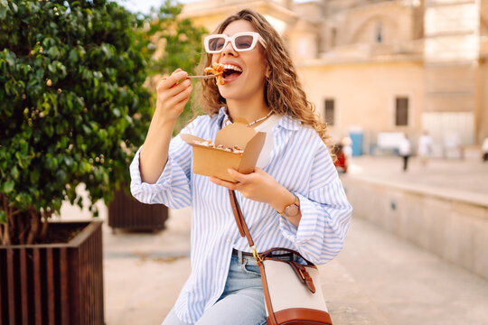 Fototapeta Smiling tourist eats street food from a takeaway box on sunny city streets. Young woman tries Italian pasta in the open air. Food concept, urban, tourism.
