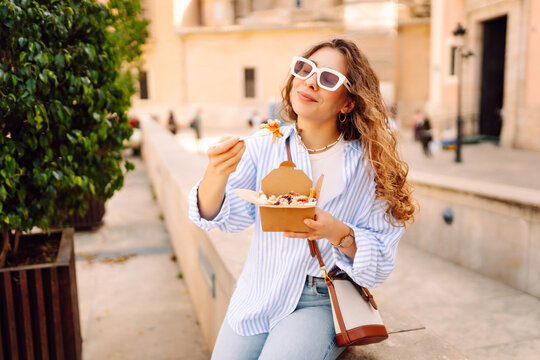 Fototapeta Smiling tourist eats street food from a takeaway box on sunny city streets. Young woman tries Italian pasta in the open air. Food concept, urban, tourism.
