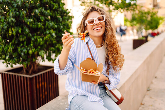 Fototapeta Smiling tourist eats street food from a takeaway box on sunny city streets. Young woman tries Italian pasta in the open air. Food concept, urban, tourism.