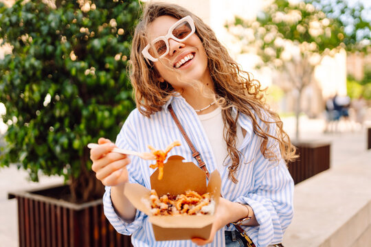 Fototapeta Smiling tourist eats street food from a takeaway box on sunny city streets. Young woman tries Italian pasta in the open air. Food concept, urban, tourism.