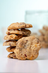 A vertical close-up shot of a stack of homemade chocolate chip and nut cookies, with a single cookie leaning against the stack on a white surface, with a blurred background.
