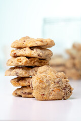 A vertical close-up shot of a stack of homemade chocolate chip and nut cookies, with a single cookie leaning against the stack on a white surface, with a blurred background.