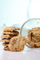 A vertical close-up shot of a stack of homemade chocolate chip and nut cookies, with a single cookie leaning against the stack on a white surface, with a blurred background.