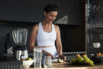 Middle aged Caucasian woman preparing healthy meal in modern kitchen, slicing green apple on wooden board near blender and fresh fruits, wearing sportswear, focused on task