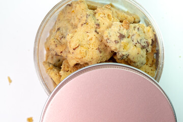 A top-down, close-up shot of a transparent jar filled with almond cookies. The cookies appear crunchy, with visible almond pieces. The rose gold-colored lid of the jar is placed in the foreground.