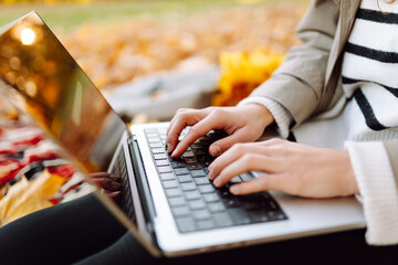 Beautiful female freelancer in raincoat and hat with laptop in meadow with fallen leaves at sunset. Nice woman works in an autumn park with laptop on sunny day. Remote work concept, education.