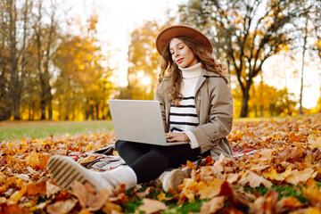 Beautiful female freelancer in raincoat and hat with laptop in meadow with fallen leaves at sunset....