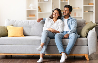 Joyful Indian couple, comfortably sitting on a sofa in their homey space, sharing a light, dreamy...