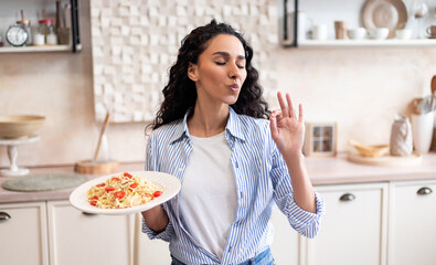 Young latin housewife tasting homemade pasta, gesturing and approving taste with closed eyes,...