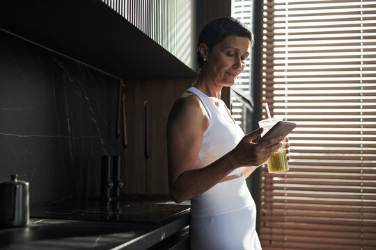 Middle aged Caucasian woman standing in modern kitchen holding smartphone in one hand and drinking green smoothie from cup with straw - Powered by Adobe