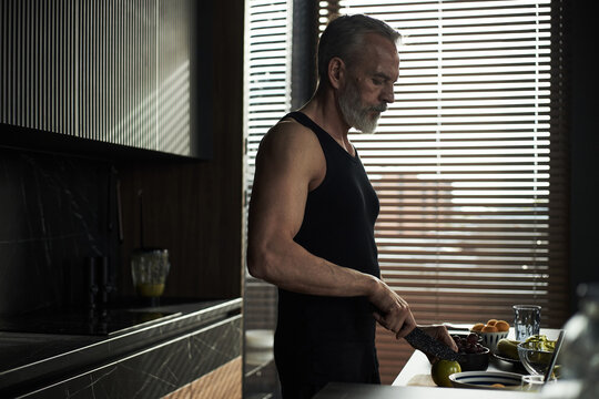 Middle aged Caucasian man preparing food in modern kitchen, slicing ingredients on countertop with knife, focused expression, natural light streaming through window blinds