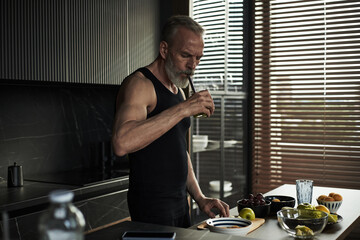 Middle aged Caucasian man standing in modern kitchen preparing healthy breakfast, drinking smoothie, surrounded by fresh fruits and vegetables on countertop