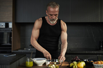 Caucasian middle aged man preparing food in modern kitchen, slicing fresh fruit on wooden countertop, showing muscular arms and focused expression while working with ingredients