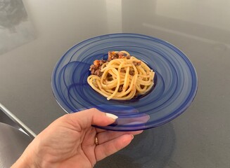 Woman holding a plate of square shaped spaghetti topped with a bolognese sauce in a glass blue bowl against a grey background 