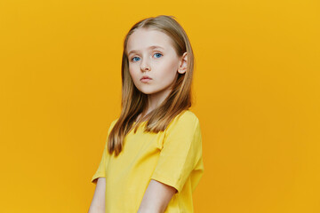 A young girl with long blonde hair stands against a bright yellow background, wearing a yellow...