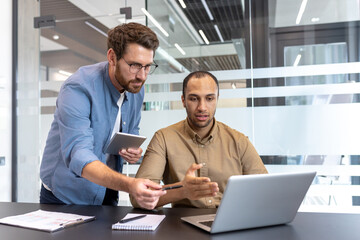 Two colleagues discuss work in a contemporary office, utilizing a laptop and a tablet.