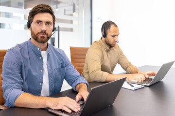 Two office professionals using laptops, wearing headsets, and engaged in a focused environment....