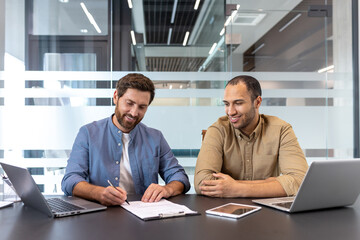 Two professionals are collaborating in an office space. One is signing papers while the other...