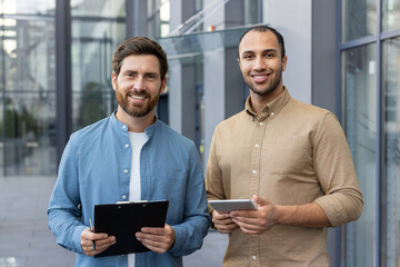 Two men stand outside a modern office building. They hold a clipboard and tablet, smiling professionally, representing teamwork or professional collaboration in a business environment.