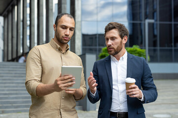 Two individuals engage in professional discussion looking at a tablet outdoors. The environment...