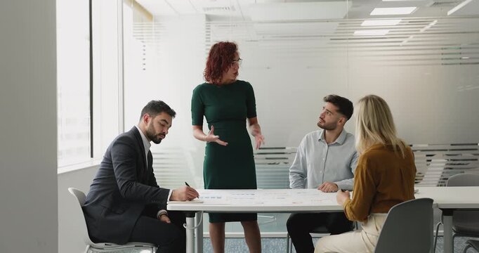Professional woman addressing group of engaged colleagues or shareholders seated around table, presenting business idea, explains performance data shown in printed charts spread out on desk. Seminar
