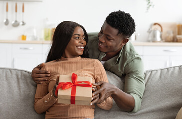 Loving african american man hugging his wife, giving gift box, making great surprise for St. Valentines Day, home interior, free space. Happy black guy embracing his girlfriend, holding present