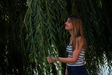 Young woman standing near a weeping willow, surrounded by foliage, gently leaning in as if inhaling...