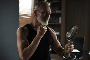 Middle aged Caucasian man smiling while holding mirror and touching chin, standing in modern room...