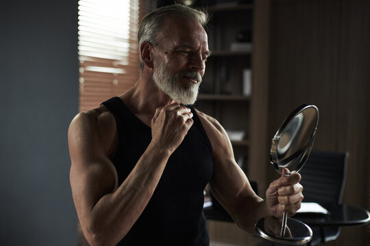 Middle aged Caucasian man smiling while holding mirror and touching chin, standing in modern room with muscular arms visible, showing confident grooming or self care routine