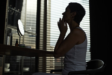 Middle aged Caucasian woman sitting at table touching face while looking into mirror, applying skincare product in modern room with window blinds in background