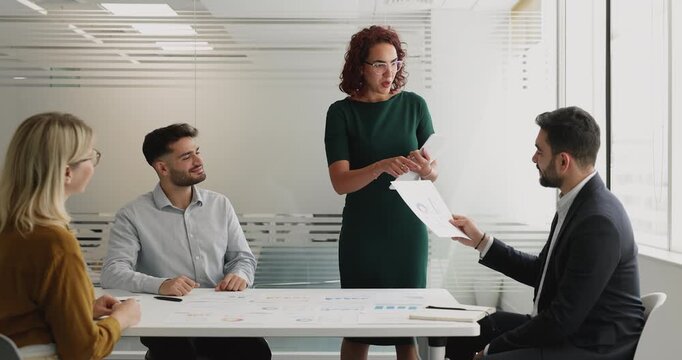 Middle-aged 40s businesswoman, project leader or presenter hands out printed charts and reports to colleagues or shareholders during formal meeting in modern conference room. Data-driven consulting