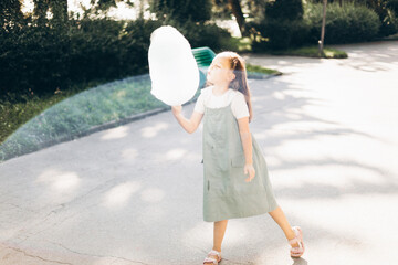 A young girl with long brown hair enjoying a sunny day, holding a large cotton candy in a green...