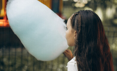 A young girl with long hair enjoys a fluffy blue cotton candy at an outdoor festival, surrounded by...