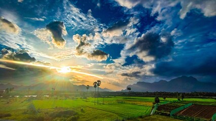 Sunrise over lush green fields in Aralvaimozhi, Kanyakumari, with dramatic clouds, distant...