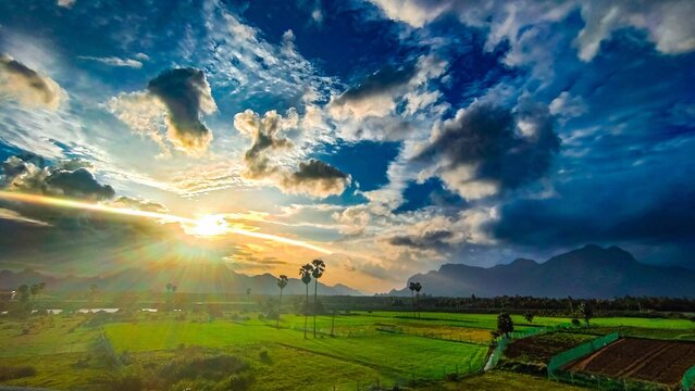 Sunrise over lush green fields in Aralvaimozhi, Kanyakumari, with dramatic clouds, distant mountains, and palm trees creating a stunning natural landscape during dawn time 