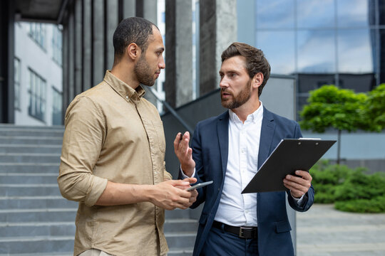 Two individuals are engaged in a professional discussion outside with a smartphone and a clipboard, exemplifying business collaboration, communication, and teamwork.