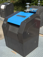 Modern black and blue recycling bin positioned on urban pavement, surrounded by greenery, promoting...