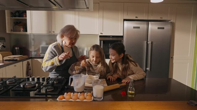 Grandma stirs with a spoon in a bowl and lets her granddaughters try while she pours water and talks