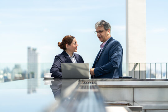 Businessman and businesswoman conversation in outdoors with laptop and coffee on rooftop terrace, cityscape in the background. - Powered by Adobe