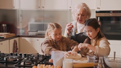A grandmother talks and watches her two granddaughters knead dough while standing next to her at the table