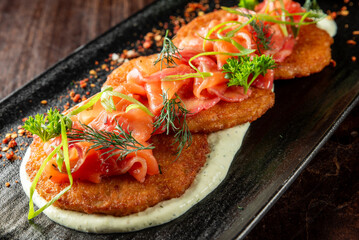 Crispy potato pancakes topped with smoked salmon, fresh herbs, and green onion on creamy dill sauce, served on a black plate.