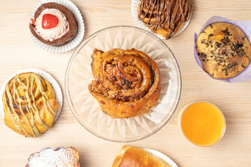 Overhead view of a wooden table laden with an assortment of freshly baked pastries, including cinnamon rolls, muffins, and a swiss roll, accompanied by a glass of refreshing orange juice