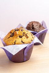 Two freshly baked muffins, one with chocolate chips and one chocolate flavored, sit in purple paper cups on a wooden table