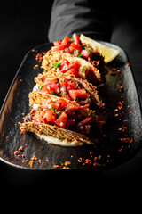 Close-up of three crispy tacos with shredded meat, fresh salsa, chili flakes, and lemon wedge on a black plate over dark background.