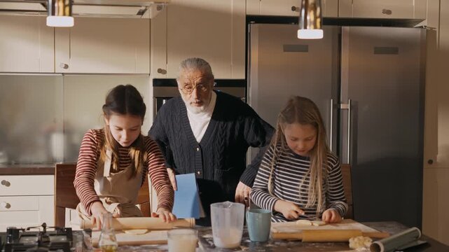 Grandpa walks into the kitchen and holds a folder and then watches as the two granddaughters cut and roll out the dough